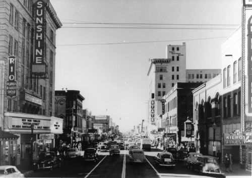 Monochrome photo of a four lane street with tall buildings on either side. Large sign saying Sunshine with movie postings below.