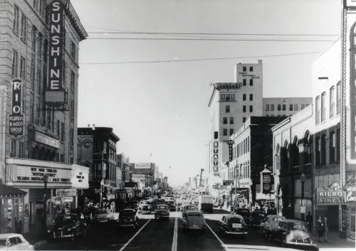 Monochrome photo of a four lane street with tall buildings on either side. Large sign saying Sunshine with movie postings below.