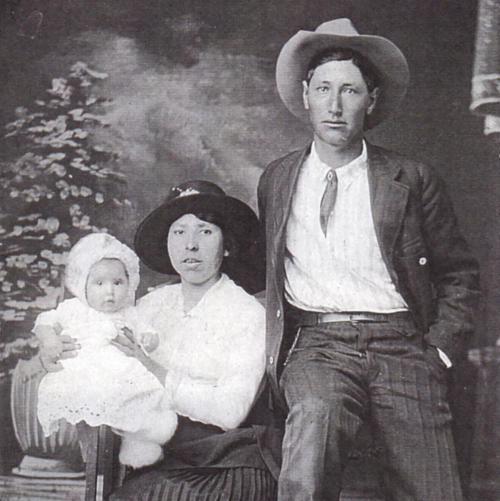 Studio posed monochrome family portrait. Man and woman in western wear. Woman holding child in gown. 
