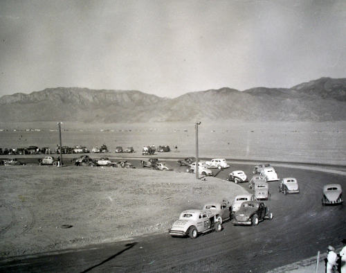 Monochrome photo of a racetrack with desert and mountains beyond. Cars have the old style curved trunk area.