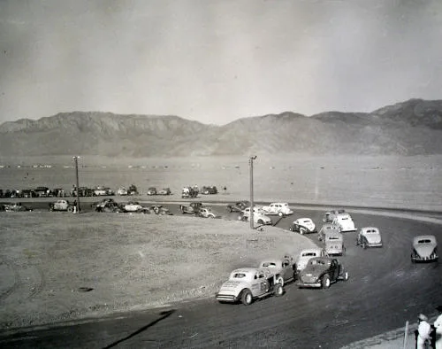 Monochrome photo of a racetrack with desert and mountains beyond. Cars have the old style curved trunk area.