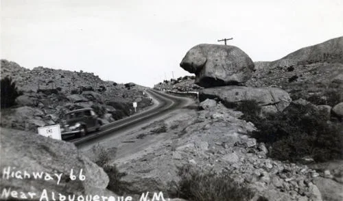 Monochrome photo from behind large rocks next to a two lane road. Written on top: Highway 66 Near Albuquerque N.M.